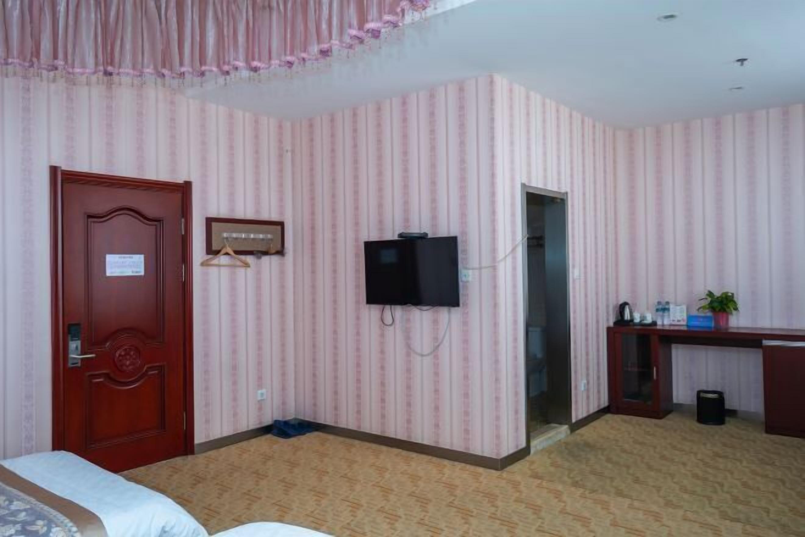 A hotel room with striped wallpaper, a wooden door, a television mounted on the wall, a desk with a computer, and a small trash bin.
