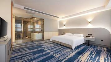 Una habitación de hotel moderna con una cama grande, un televisor de pantalla plana, un box de ducha de vidrio y una alfombra azul estampada.