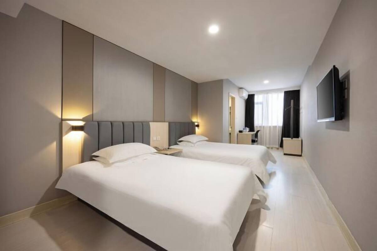 A hotel room with two beds, a TV mounted on the wall, and a bedside table with a lamp.