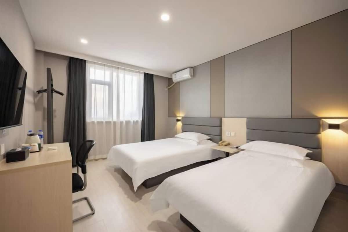 A hotel room with two beds, a desk, a chair, a TV, and an air conditioning unit.