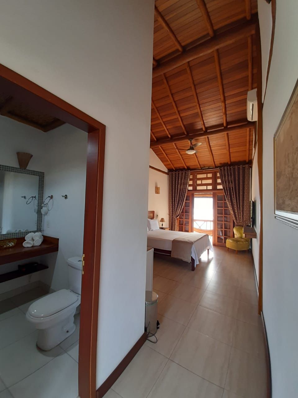 Quarto de hotel com teto de madeira, banheiro com vaso sanitário e pia, uma cama e uma pequena cadeira amarela.