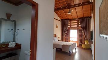 Quarto de hotel com teto de madeira, banheiro com vaso sanitário e pia, uma cama e uma pequena cadeira amarela.
