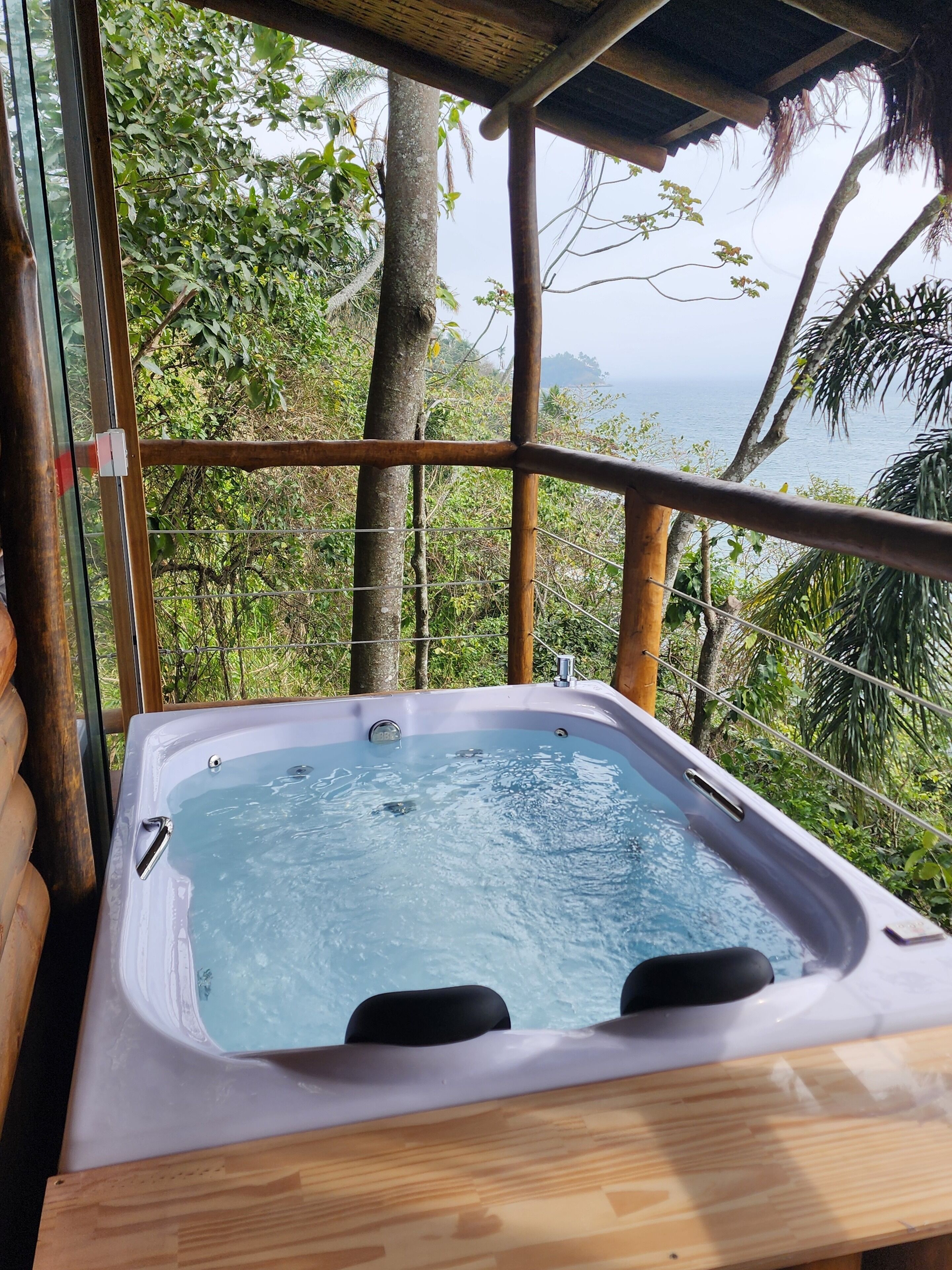 A hot tub with a wooden deck, surrounded by a bamboo fence, overlooking a lush landscape and the sea.