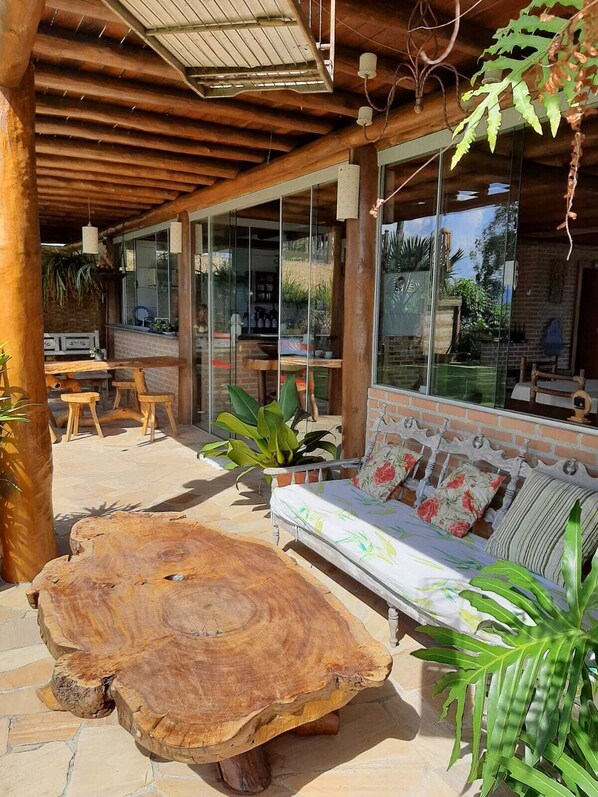 An outdoor patio area with a wooden bench, a large wooden table, and a sofa with floral cushions. - Gidu House (Ilhabela)