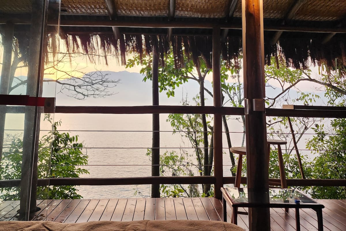 Una habitación con terraza de madera, techo de paja y vista hacia el agua.