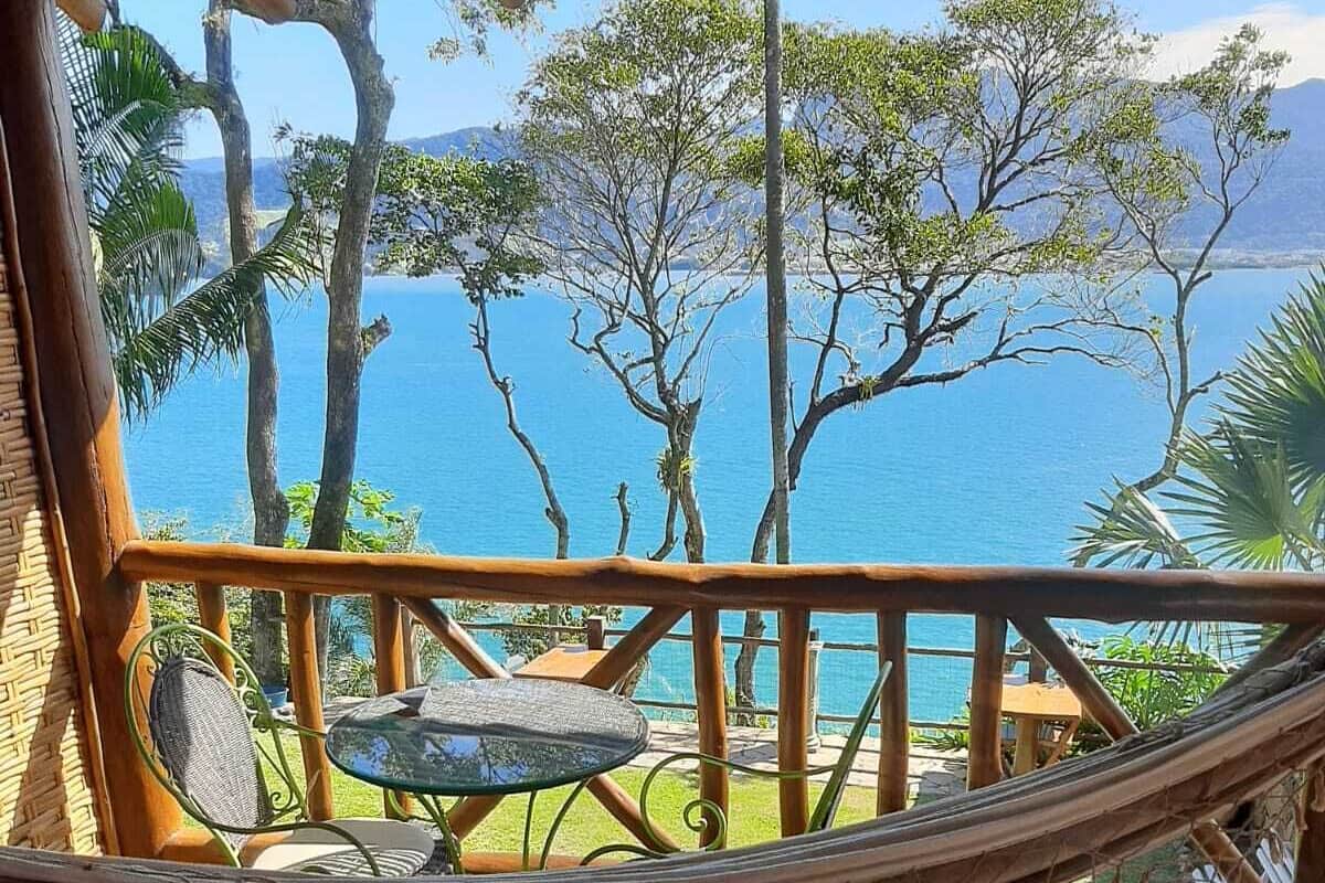 Una terraza de madera con una hamaca, una pequeña mesa redonda y una silla, con vistas a un cuerpo de agua y árboles.