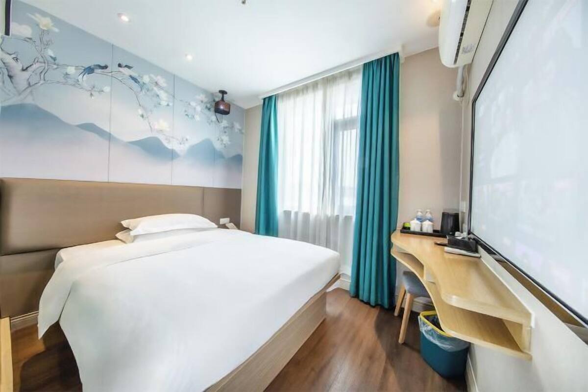 A hotel room with a bed, a desk, a chair, a waste bin, and a large window with curtains.