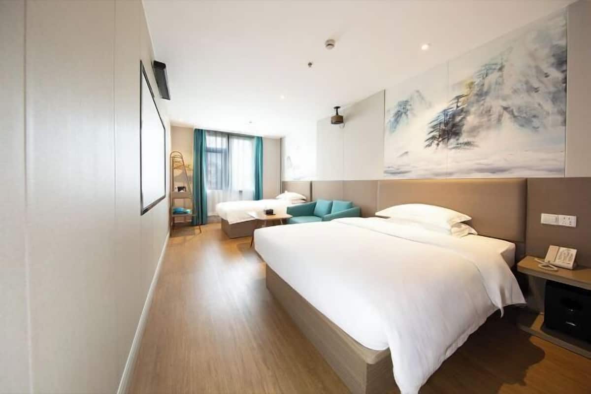 A modern hotel room with a large bed, a seating area with a sofa and armchair, a painting on the wall, and a bedside table with a lamp.