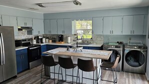 A modern kitchen with a central island, bar stools, a built-in washing machine, and a large window providing natural light.