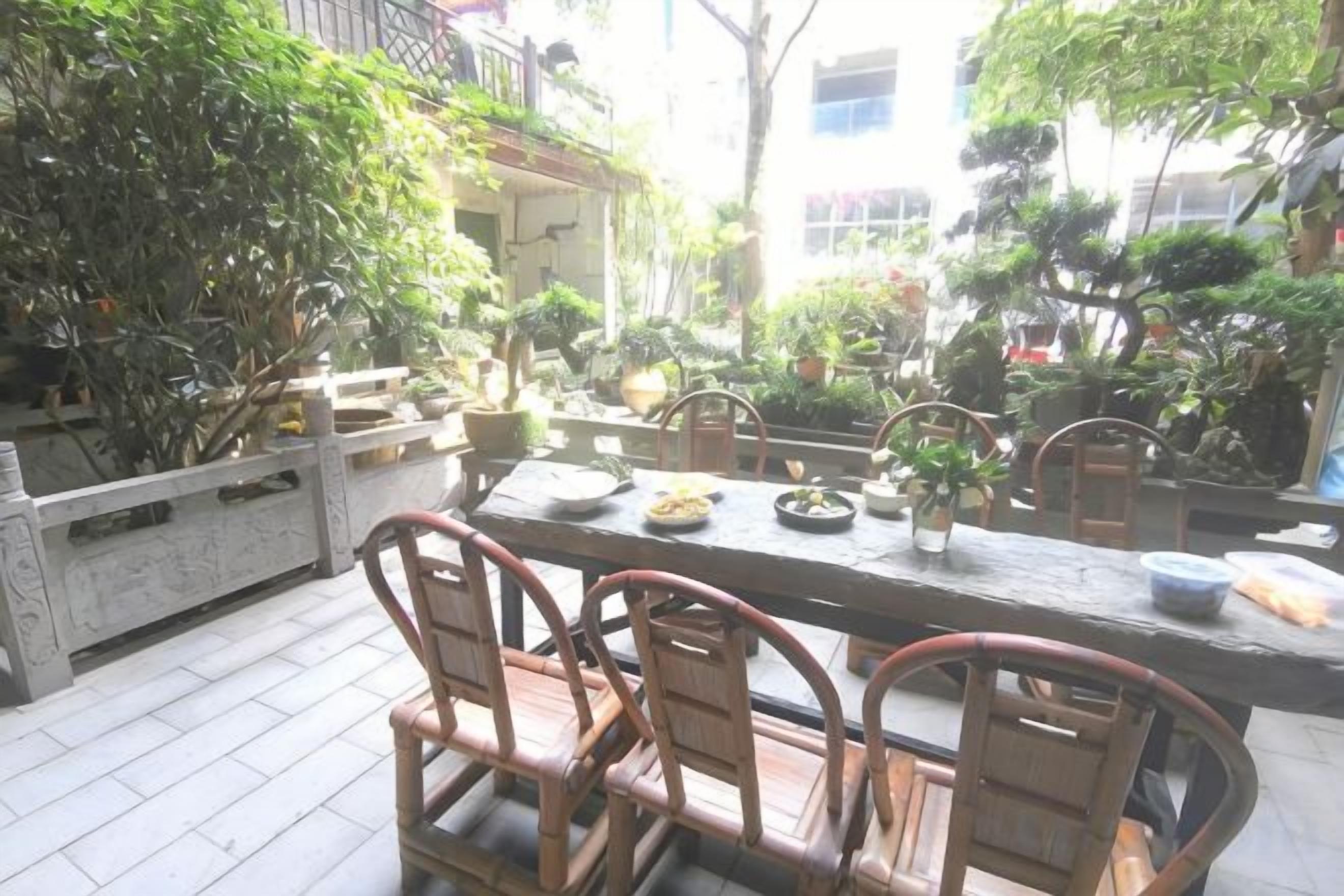 An outdoor dining area with a long table and chairs, surrounded by potted plants and trees.