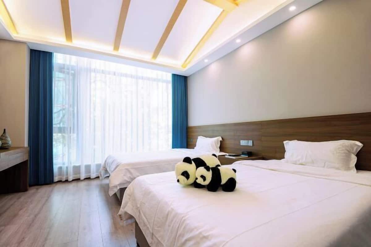 Habitación de hotel con dos camas, cabecero de madera, mesita de noche y un amplio ventanal con cortinas.
