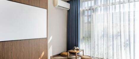 A modern hotel room with a large window, a flat-screen TV, and a small table with a lamp.