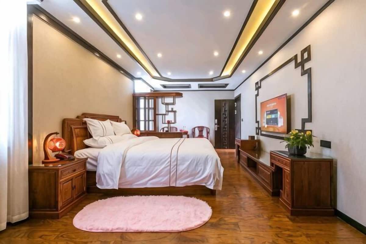 Un dormitorio con cama, mesitas de noche, escritorio, silla, espejo, televisión montada en la pared, suelo de madera y alfombra rosa.
