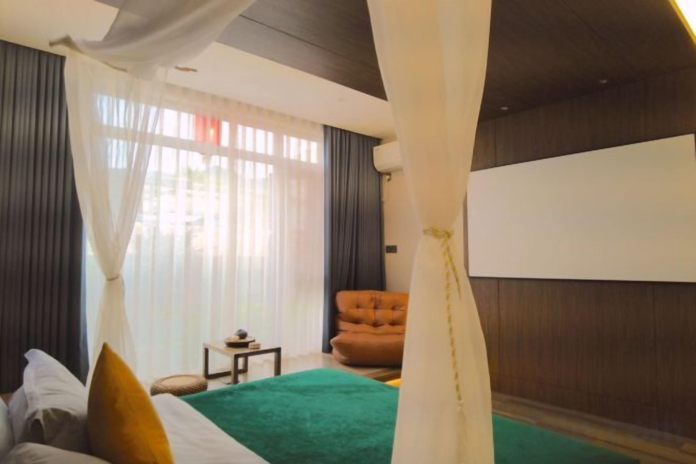 Quarto com cama, cadeira, mesinha e uma janela grande com cortinas.
