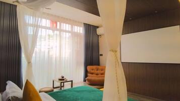 Quarto com cama, cadeira, mesinha e uma janela grande com cortinas.