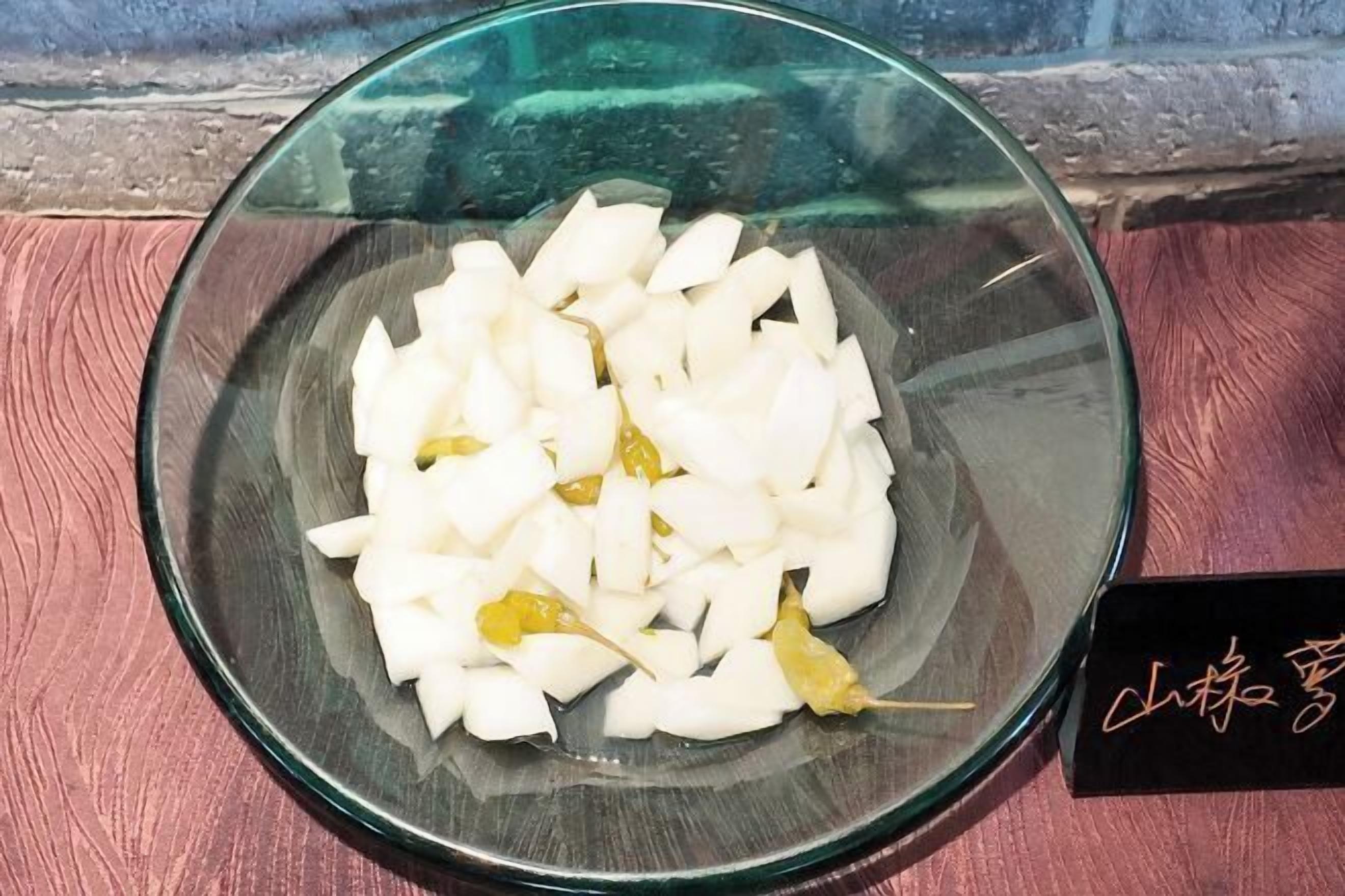 A glass bowl containing sliced white fruit and a few yellow peppers on a wooden surface.
