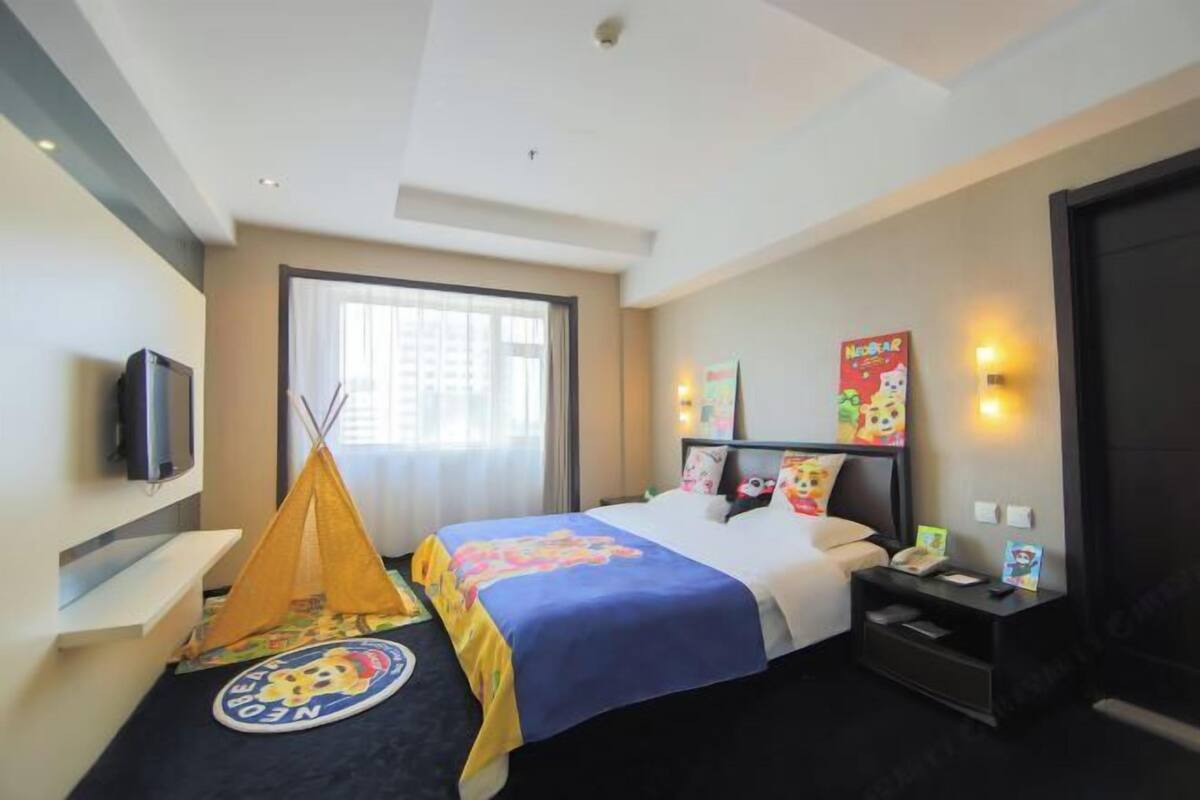 A hotel room with a bed, a television, a window, a nightstand, and a wall-mounted light.