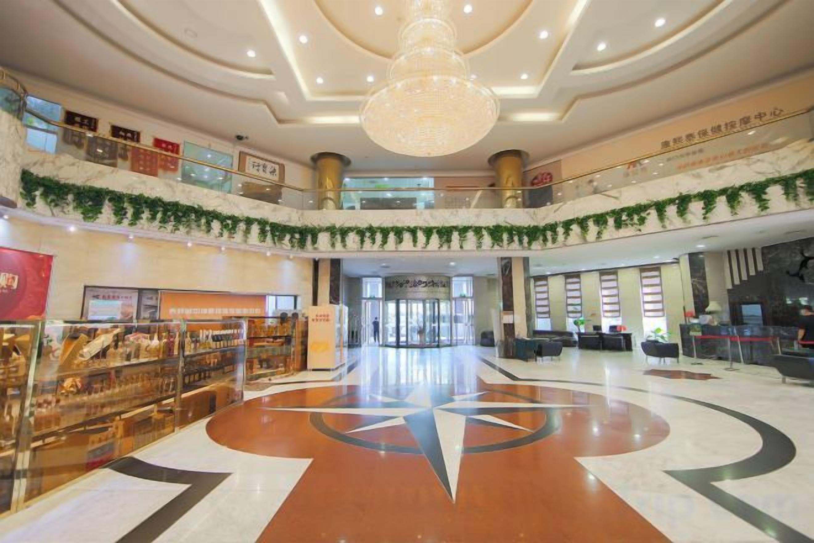 A spacious lobby with a central geometric floor design, a large chandelier, and a second-floor balcony with greenery.