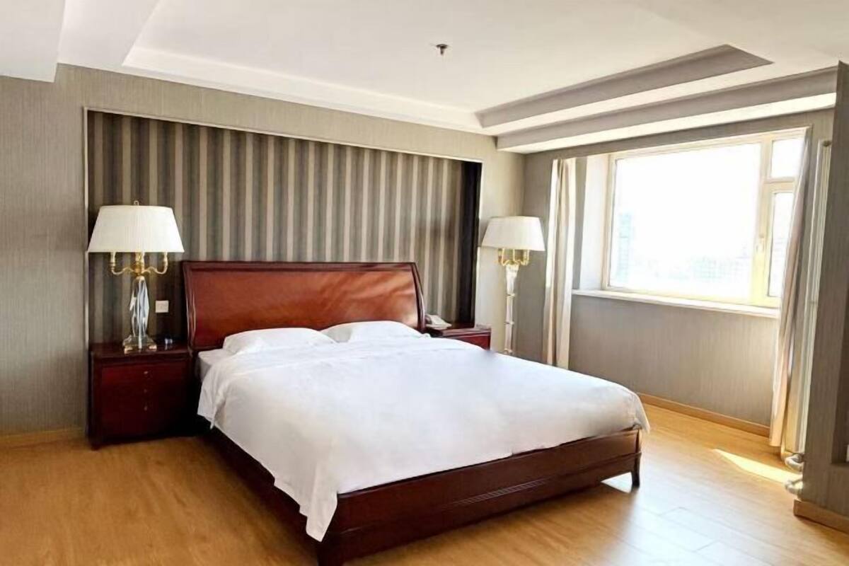 A hotel room with a large bed, two bedside tables with lamps, a window with curtains, and a striped headboard.