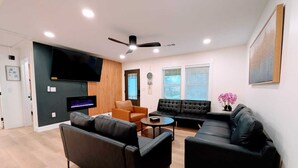 A modern living room with a black sofa, a brown armchair, and a black couch. There is a flat-screen TV mounted on the wall, a ceiling fan, and a small table with a vase of flowers. - 10 Burnett Home (Rome)