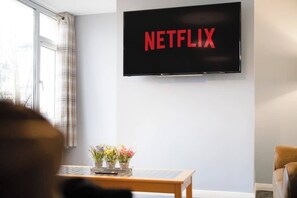 A room with a television displaying 'NETFLIX,' a window with curtains, and a small table with flowers. - 3BR in Richmond sleeps 5, Garden and Free Parking (Colburn)