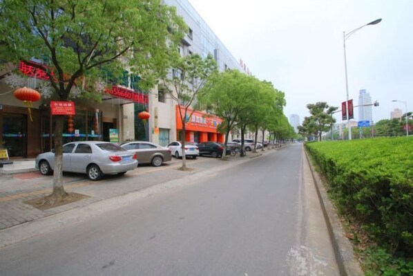 A street lined with parked cars and storefronts, with trees and a hedge along the sidewalk. - Yihao Hotel (Shanghai Huinan subway station store) (Shanghai)