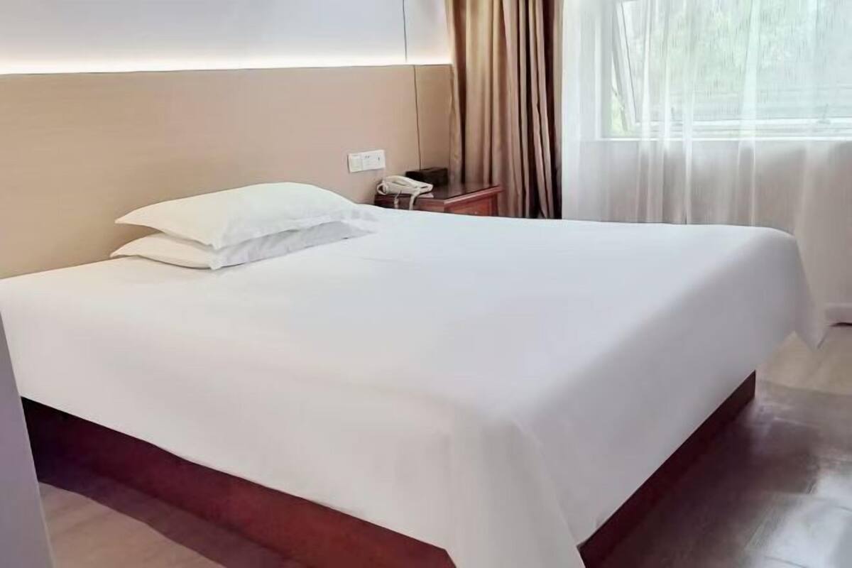 A neatly made bed with white linens, a bedside telephone, and a window with sheer curtains.