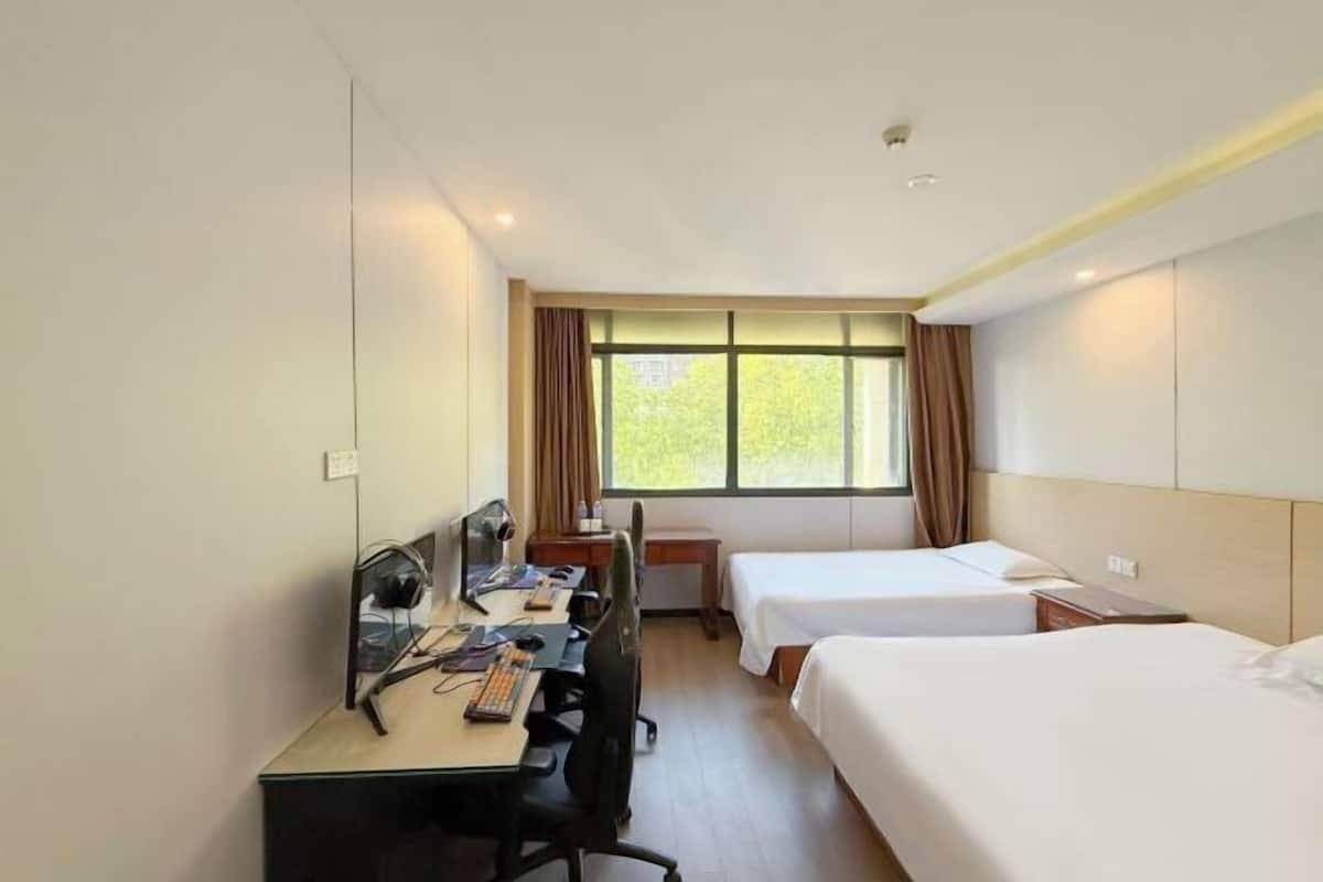 A hotel room with a desk, two beds, a window with curtains, and a view of greenery outside.