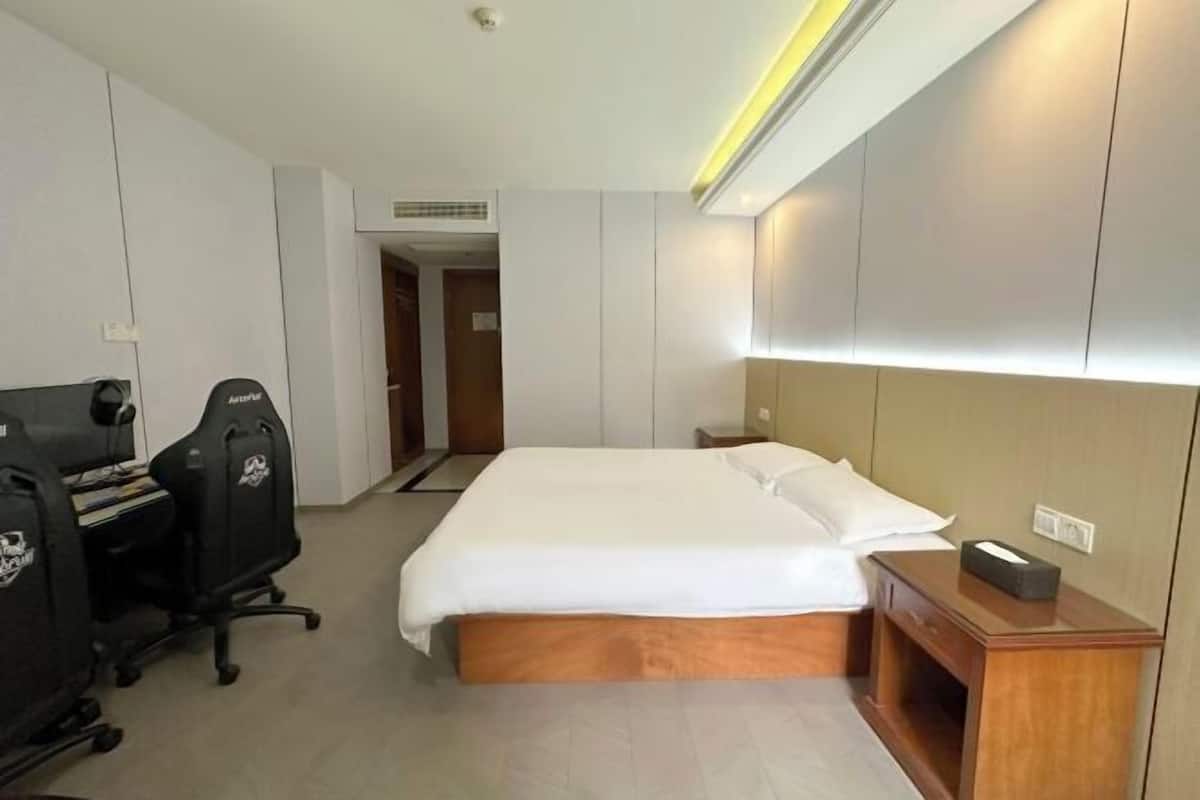A hotel room with a bed, a desk with two chairs, a bedside table, and a wall-mounted shelf.