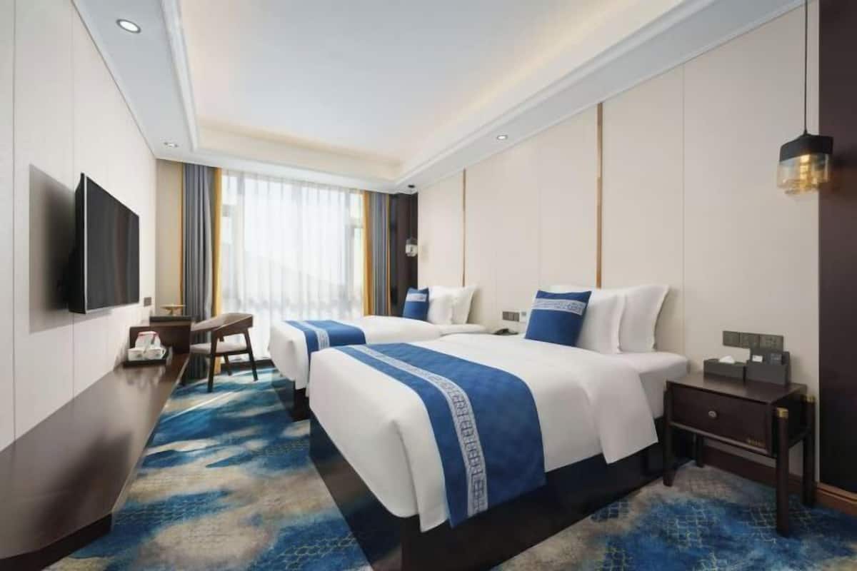 Habitación de hotel con dos camas, un escritorio, televisión y un ventanal con cortinas.
