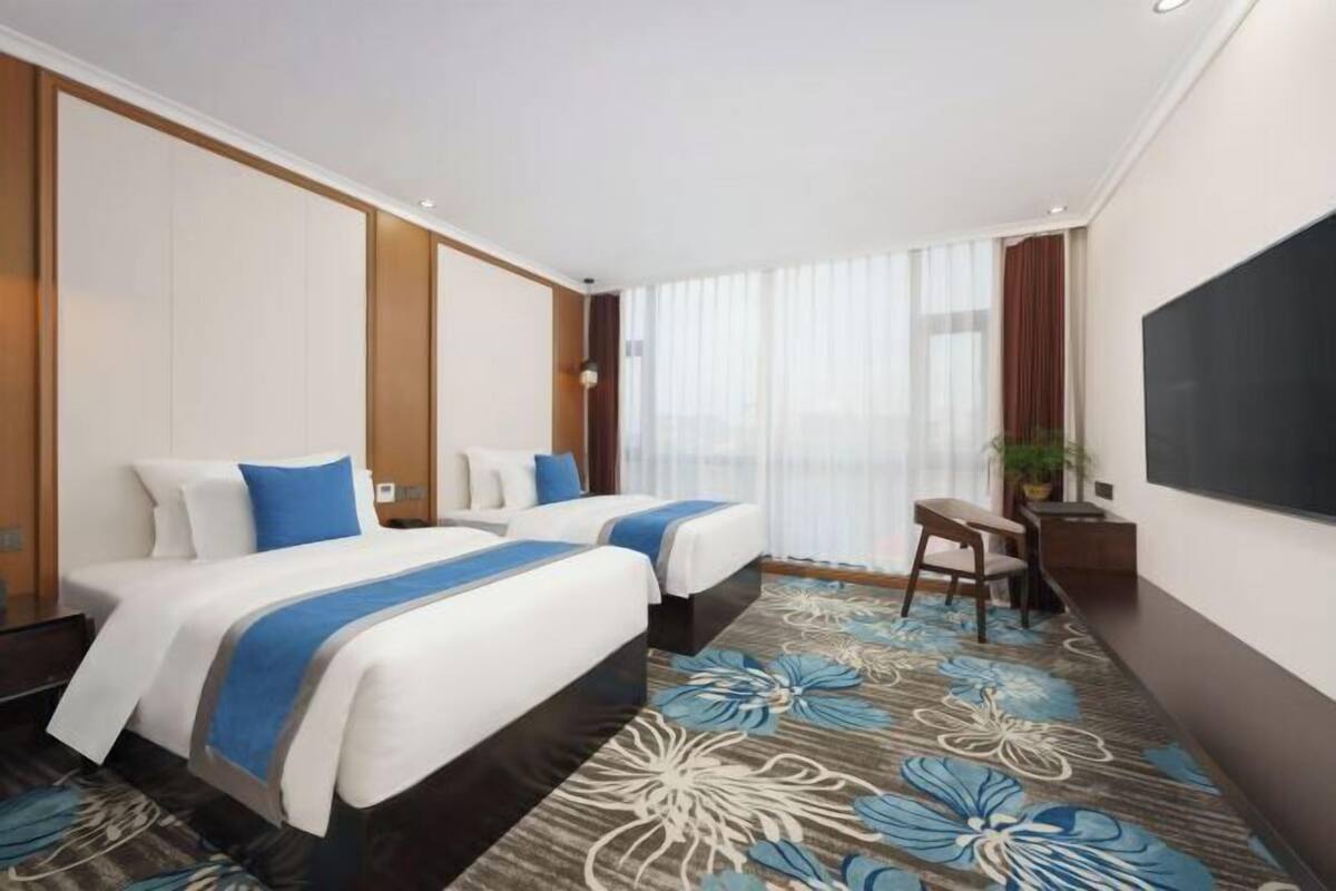Habitación de hotel con dos camas, un escritorio, una silla, un televisor y un amplio ventanal con cortinas.