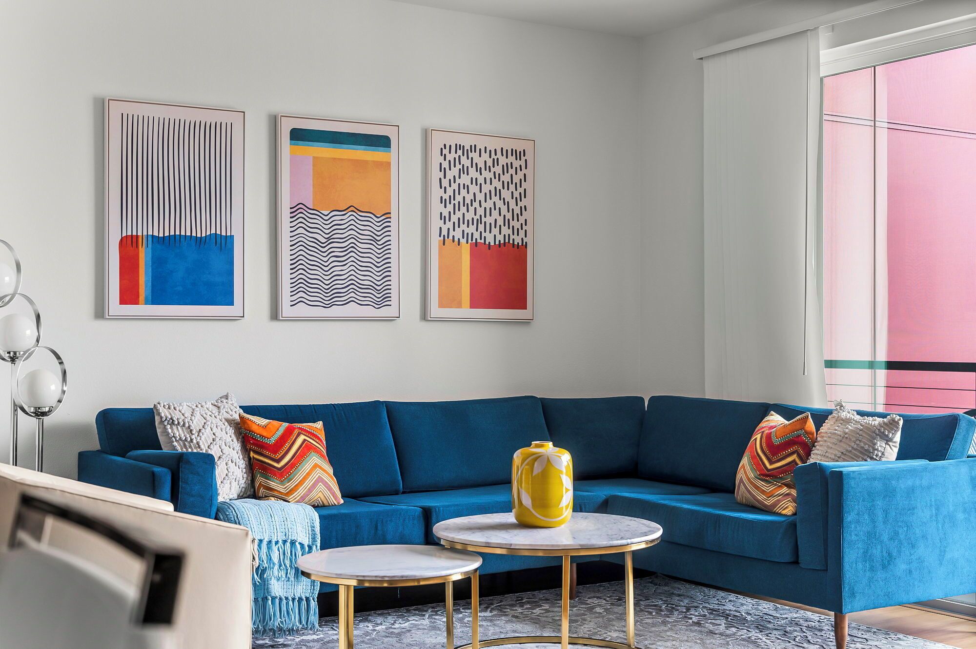 Una sala de estar moderna con un sofá seccional azul, tres obras de arte abstractas enmarcadas, un jarrón amarillo y una mesa redonda dorada.