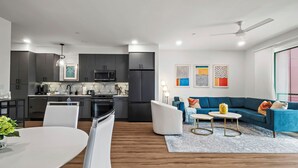 A modern kitchen with dark cabinets, a microwave, and a stove. There is a dining area with a white table and chairs. A blue sofa and a small round table are in the living area. - The Mercer (New Braunfels)