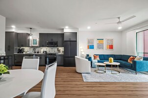 Cozinha moderna com armários escuros, micro-ondas e fogão. Possui uma área de jantar com mesa e cadeiras brancas. Um sofá azul e uma pequena mesa redonda estão na sala.