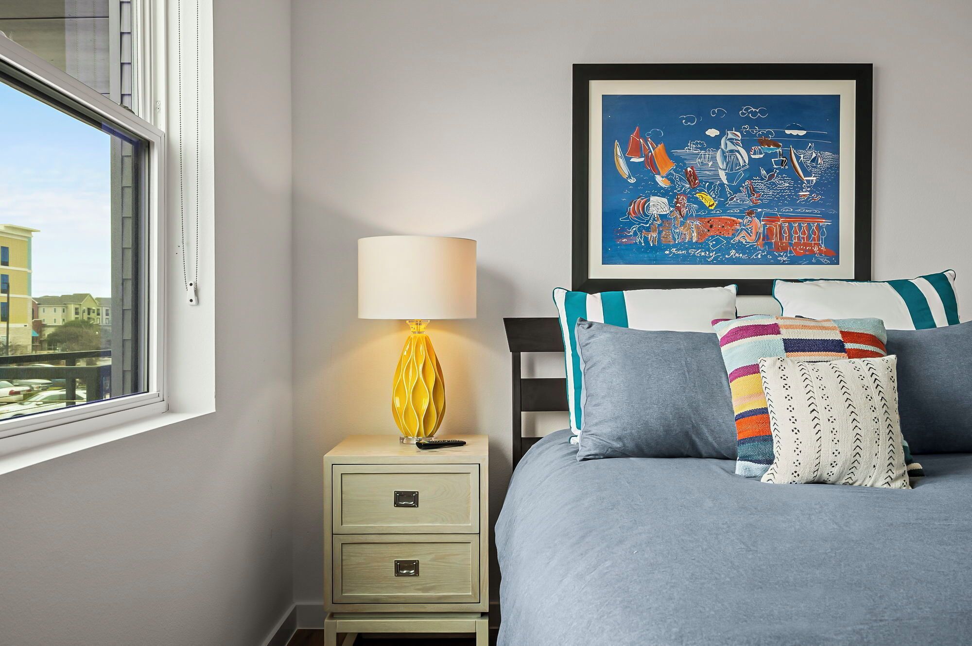 A bedroom with a bed, a bedside table with a lamp, a framed artwork, and a window with a view of buildings.