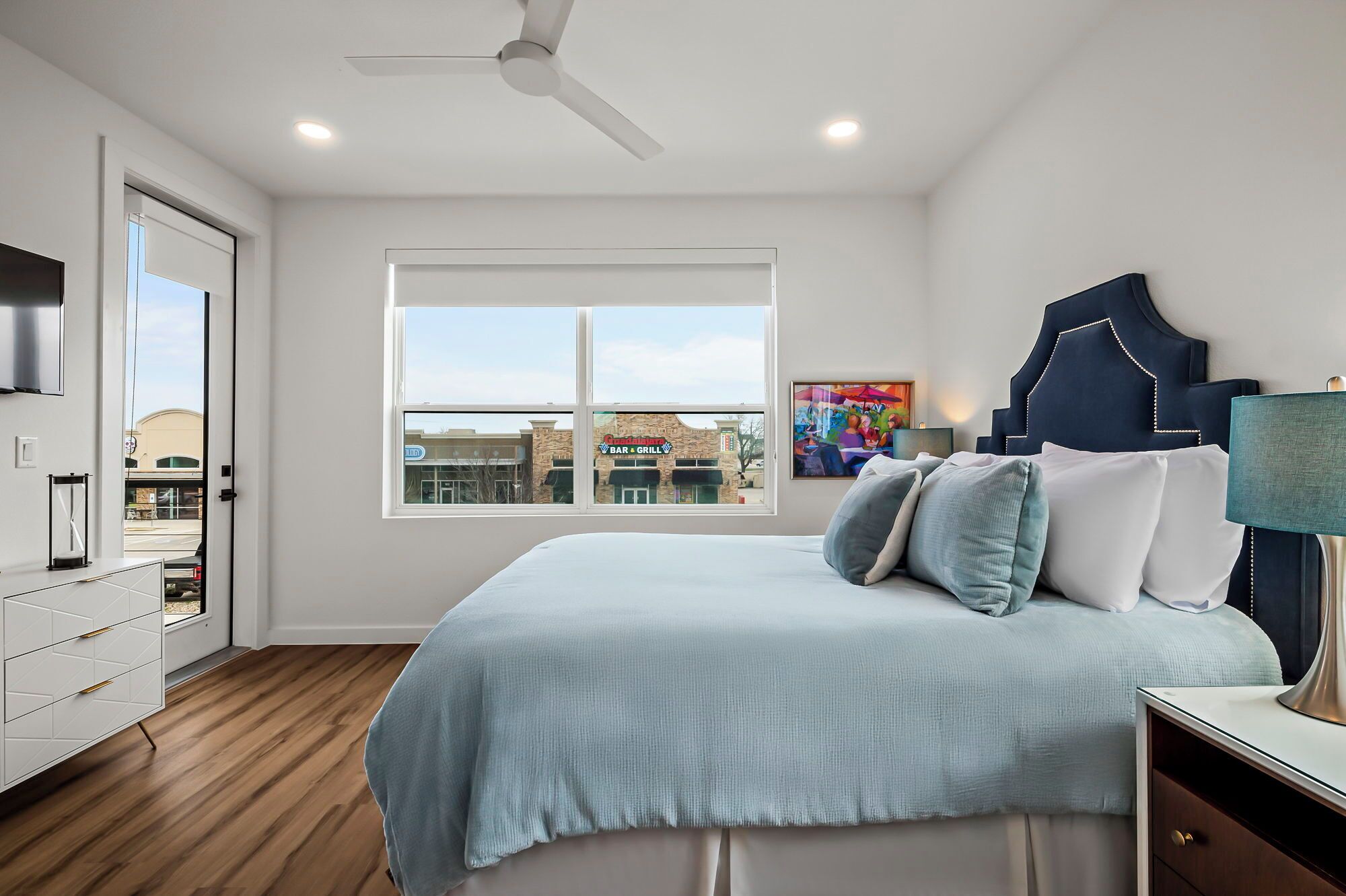 A bedroom with a bed, a ceiling fan, a window with a view of buildings, and a TV on the wall.
