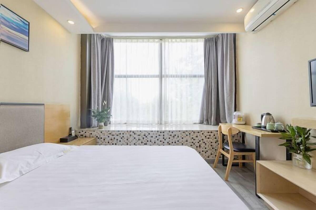 A hotel room with a bed, a desk with a chair, a window with curtains, a potted plant, and a wall-mounted air conditioner.