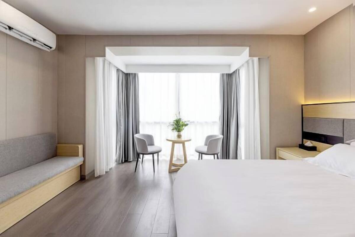 A modern hotel room with a large bed, a seating area, a small table with chairs, and a window with curtains.