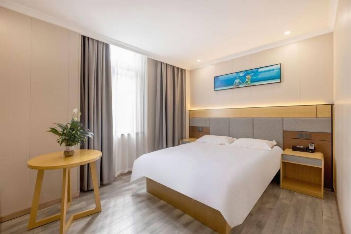 A hotel room with a bed, a bedside table with a lamp, a small table with a plant, and a painting on the wall.