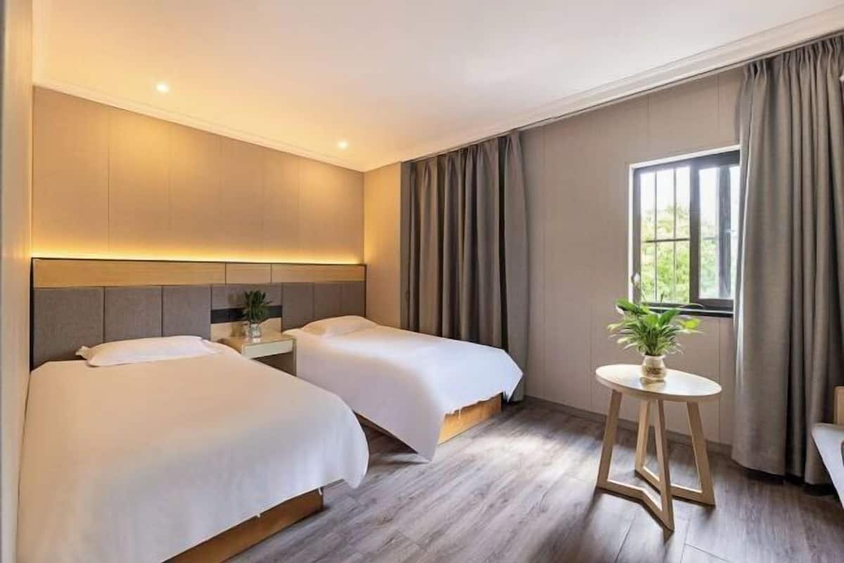 A hotel room with two beds, a bedside table with a plant, and a window with curtains.