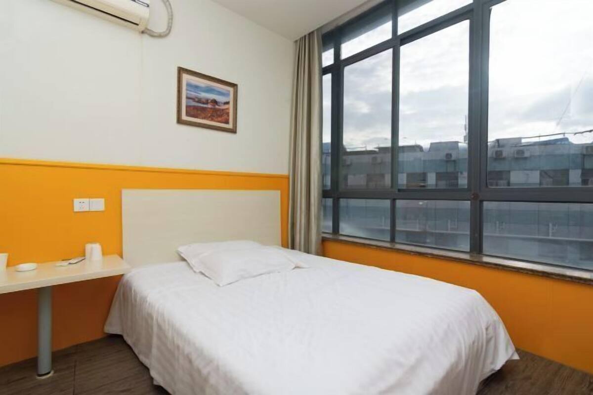 A hotel room with a bed, a window with a view of buildings, a small table with a cup, and an air conditioning unit.