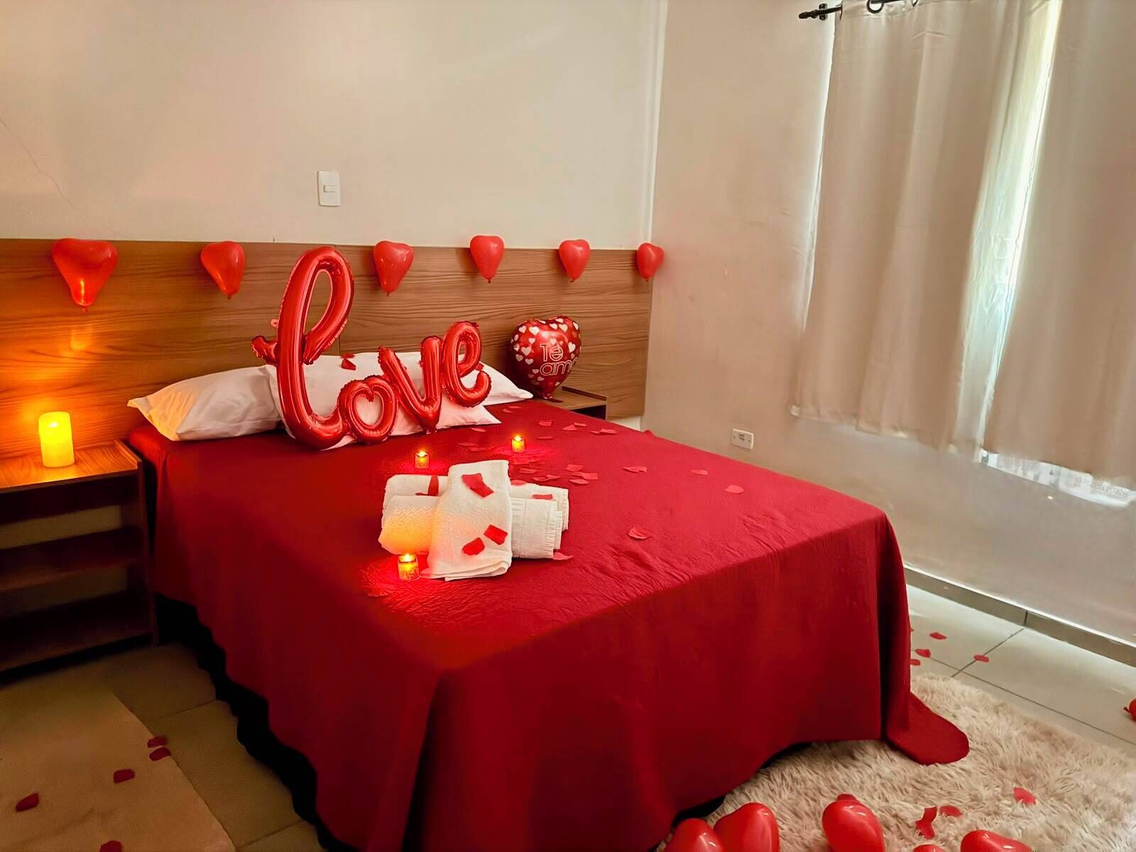 Una habitación con cama, mesita de noche, una pared decorada con corazones y una ventana con cortinas.