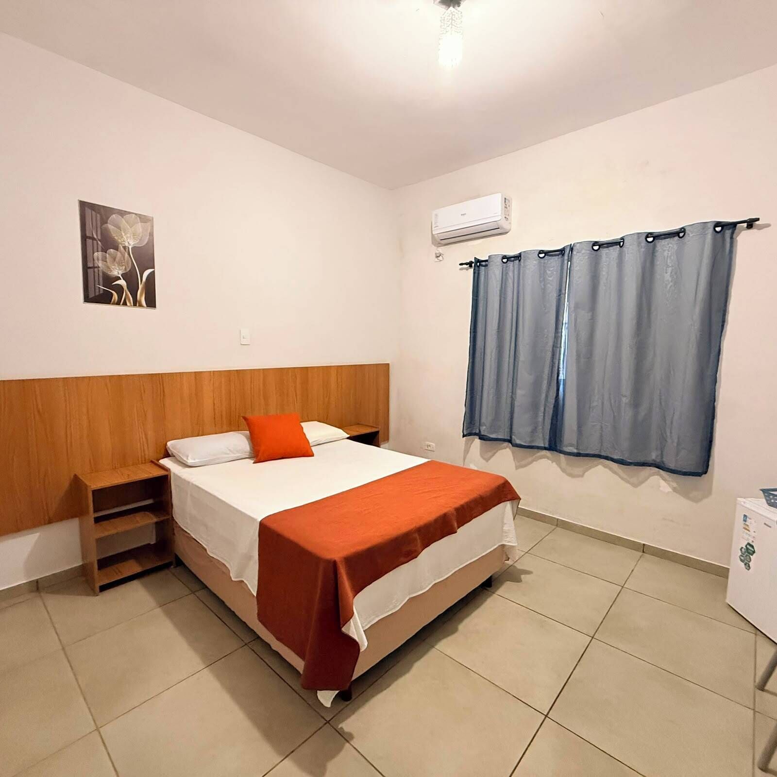 Une chambre à coucher comprenant un lit, une table de chevet, un climatiseur fixé au mur et une fenêtre avec des rideaux.