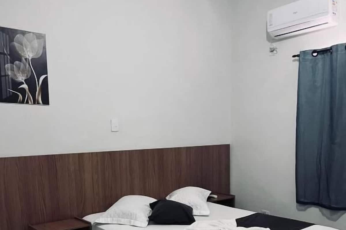 A single bed with a wooden headboard, a nightstand, a wall-mounted air conditioner, and a framed picture on the wall.