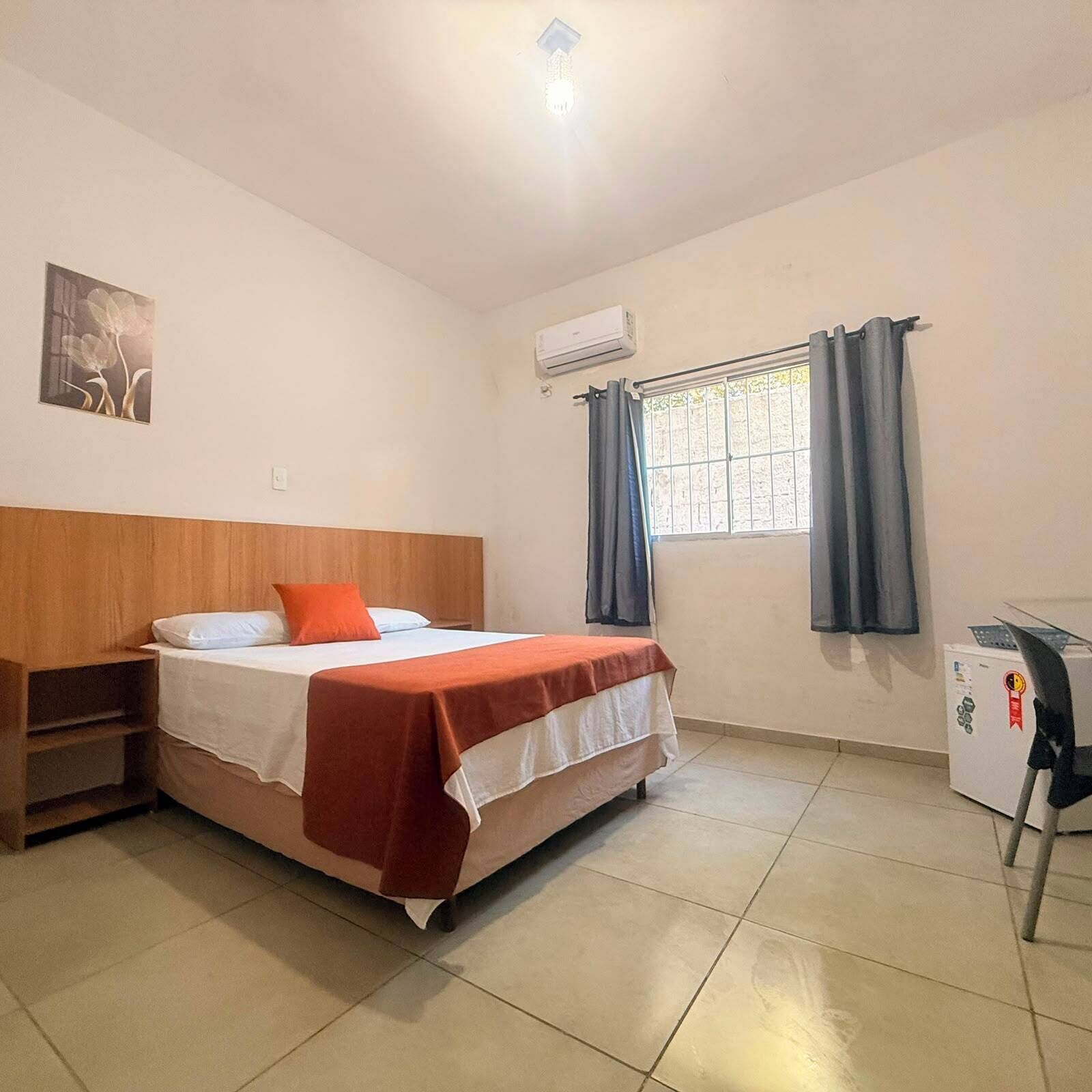 A bedroom with a bed, a nightstand, an air conditioner, a window with curtains, and a small desk with a chair.