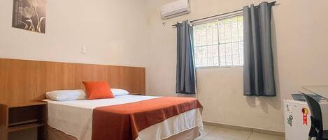 A bedroom with a bed, a nightstand, an air conditioner, a window with curtains, and a small desk with a chair.