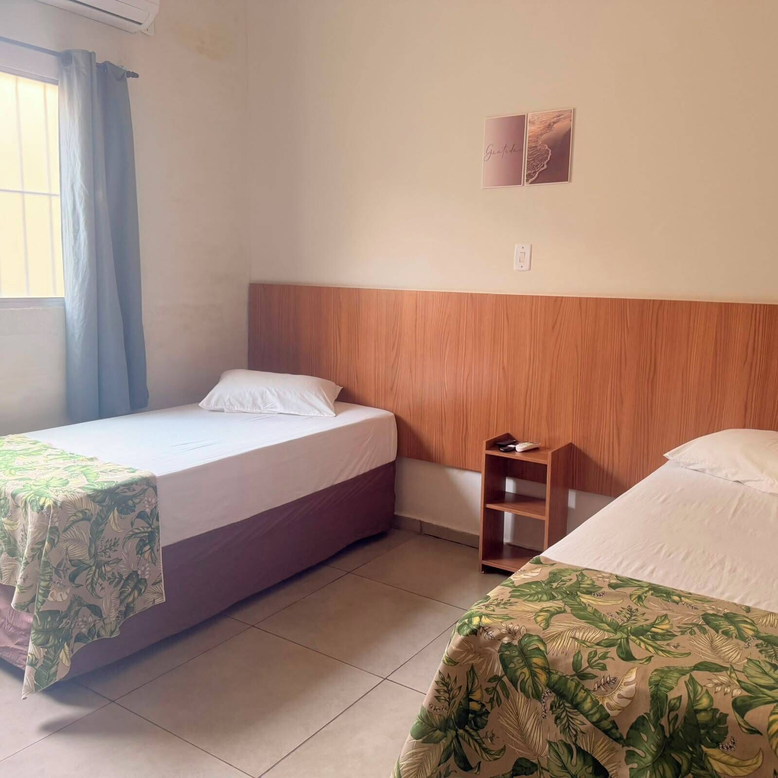 Quarto de hotel com duas camas, cabeceira de madeira, mesa de cabeceira pequena de madeira e uma janela com cortinas.