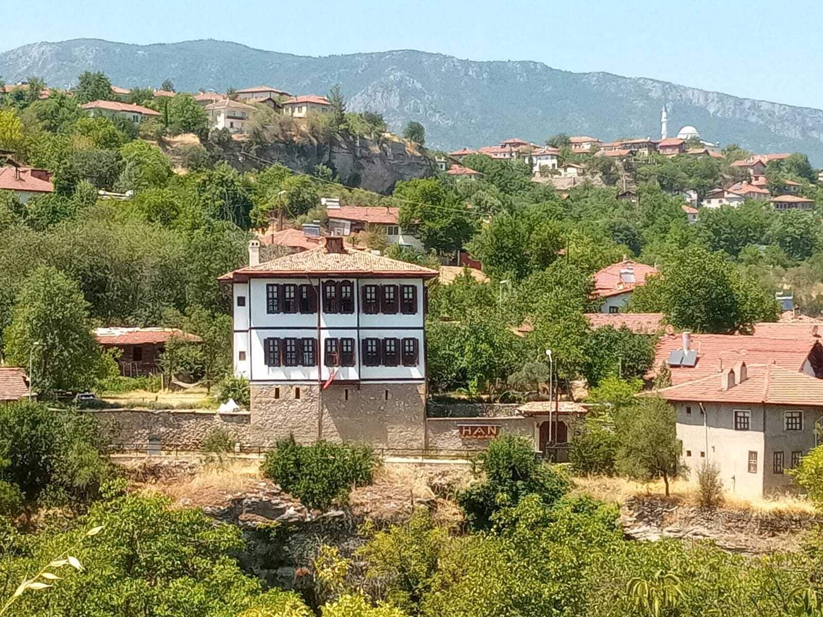 Un edificio de dos pisos con base de piedra y sección superior blanca, rodeado de vegetación y otros edificios en una zona montañosa.