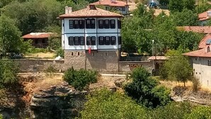 A traditional two-story building with a stone base and a tiled roof, surrounded by greenery and other residential buildings on a hillside.