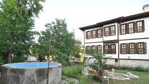 A two-story building with a white facade and brown window shutters, a concrete pool, and a garden with chairs and tables.